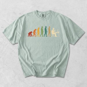 Pode incluir: Uma t-shirt de manga curta em verde s&aacute;lvia claro com uma impress&atilde;o gr&aacute;fica multicolorida que retrata a progress&atilde;o evolutiva de um primata para um humano sentado numa secret&aacute;ria de computador. O design utiliza tons de vermelho, laranja, amarelo, verde e azul.