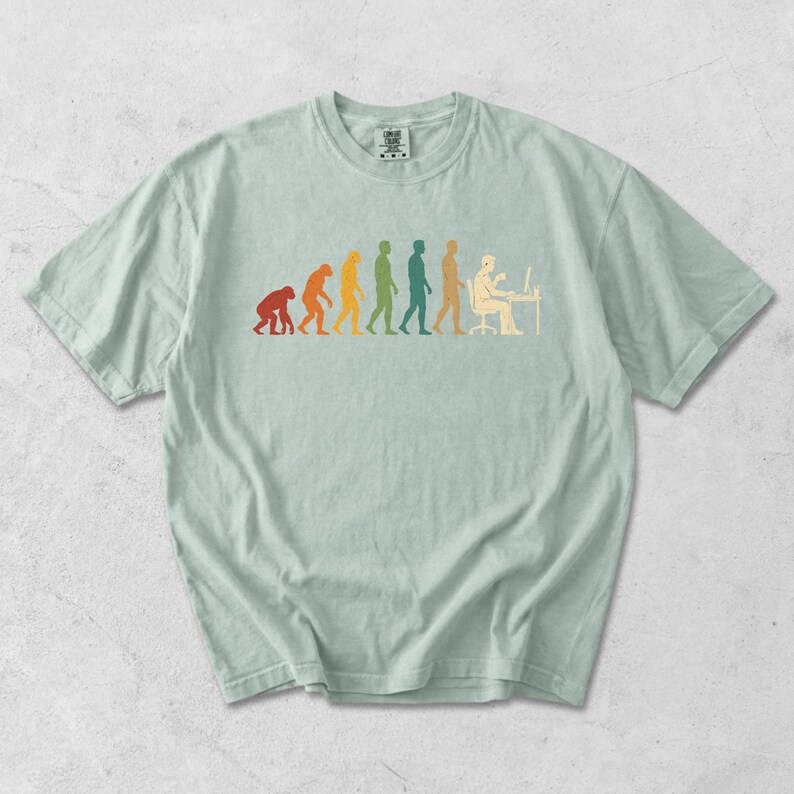 Pode incluir: Uma t-shirt de manga curta em verde s&aacute;lvia claro com uma impress&atilde;o gr&aacute;fica multicolorida que retrata a progress&atilde;o evolutiva de um primata para um humano sentado numa secret&aacute;ria de computador. O design utiliza tons de vermelho, laranja, amarelo, verde e azul.