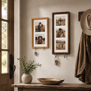 Op de afbeelding: Twee rechthoekige houten fotolijsten voor aan de muur met witte passe-partouts en geïntegreerde metalen haken aan de onderkant. De linkerlijst bevat twee foto's, de rechterlijst drie. Een sleutelbos hangt aan een van de haken.