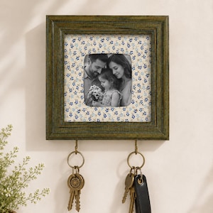 May include: A square, dark olive green wooden picture frame featuring a cream-colored mat with a small blue floral pattern. The frame holds a black and white photograph of a family and includes two brass hooks at the bottom for hanging keys.