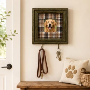 May include: A square, dark green wooden wall-mounted organizer featuring a central photo of a golden retriever against a brown and white plaid background. Two brass-colored metal hooks hang below, holding a brown leather leash and a set of keys.