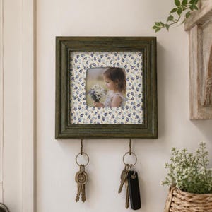 Op de afbeelding: Een vierkante, donkergroene houten fotolijst met een centrale foto van een kind dat een boeket bloemen vasthoudt. De lijst heeft een passe-partout met een blauw bloemenpatroon en twee metalen haken aan de onderkant voor sleutelbossen.