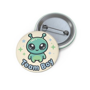 Peut inclure: Un badge rond avec épingle représentant un extraterrestre de dessin animé vert clair aux grands yeux noirs sur un fond crème. Le texte "Team Boy" est imprimé en lettres bleues en bas, entouré de petites étoiles bleues et vertes à quatre branches.