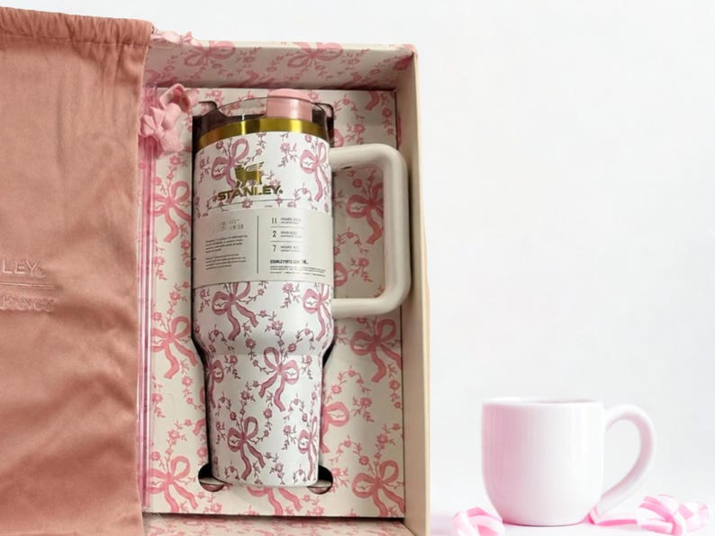 May include: A white Stanley tumbler with a pink ribbon and floral pattern, featuring a large handle and a gold-toned lid, presented in a matching patterned gift box alongside a pink velvet drawstring bag and a small, solid light pink ceramic mug.