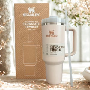 May include: A cream-colored Stanley FlowState Quencher H2.0 tumbler with a handle, a lid, and a straw, standing in front of its matching brown cardboard packaging box. The box features the Stanley logo and text reading The Quencher H2.0 FlowState Tumbler 40 oz / 1.18 L.
