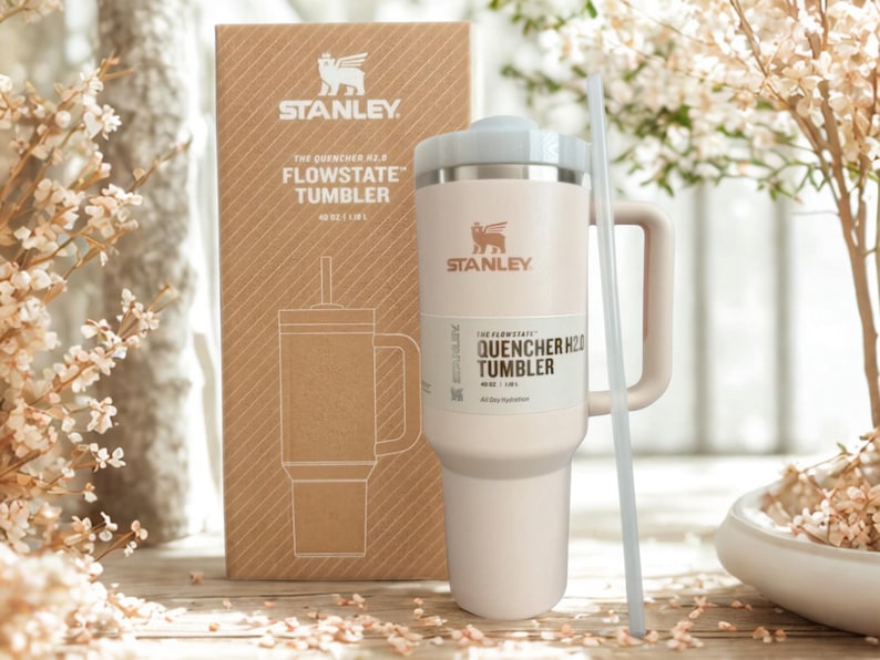 May include: A cream-colored Stanley FlowState Quencher H2.0 tumbler with a handle, a lid, and a straw, standing in front of its matching brown cardboard packaging box. The box features the Stanley logo and text reading The Quencher H2.0 FlowState Tumbler 40 oz / 1.18 L.