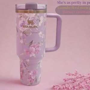 May include: A lavender Stanley tumbler with a floral pattern, a gold-toned rim, and a matching handle. The cup features the text Stanley and LoveShackFancy. The text in the image reads: She's as pretty in person, Lavender floral meets gold detail, Stanley > LoveShackFancy.