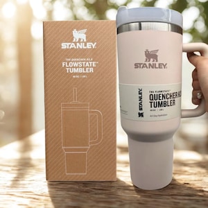 May include: A pale pink Stanley Flowstate Quencher H2.0 tumbler with a handle and lid, next to its matching brown cardboard box. Both the tumbler and the box feature the Stanley logo and the text The Flowstate Quencher Tumbler 40 oz | 1.18 L.