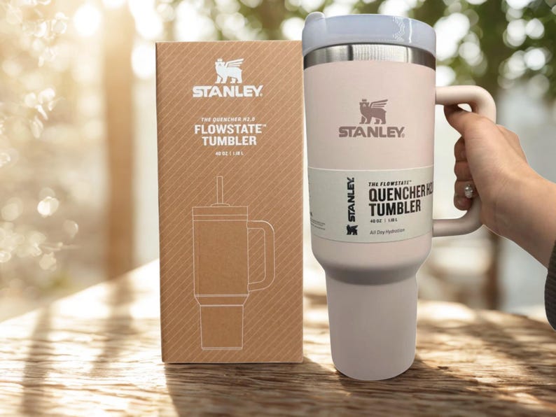 May include: A pale pink Stanley Flowstate Quencher H2.0 tumbler with a handle and lid, next to its matching brown cardboard box. Both the tumbler and the box feature the Stanley logo and the text The Flowstate Quencher Tumbler 40 oz | 1.18 L.