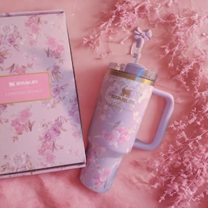 May include: A lavender Stanley tumbler with a floral pattern, a matching handle, and a lid featuring a bow-shaped straw topper. Beside it sits a rectangular box with the same floral design, featuring the Stanley and LoveShackFancy brand logos.