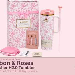 May include: A Stanley x LoveShackFancy 40 oz Quencher H2.0 tumbler featuring a white and pink ribbon and rose pattern, a matching gift box, a pink straw with a bow topper, a small pink pouch, and a promotional card showing three models in dresses.