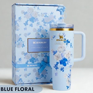 May include: A light blue Stanley tumbler with a floral pattern and a matching gift box. The tumbler features a gold rim, a handle, and a straw with a small blue bow. The Stanley and LoveShackFancy logos are printed on both the tumbler and the box.
