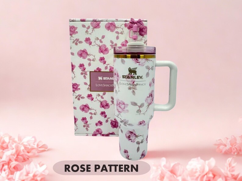 May include: A white Stanley LoveShackFancy tumbler with a pink rose pattern, a handle, and a clear lid with a pink straw topper. The tumbler is displayed in front of a matching rectangular box featuring the same floral design and brand text.