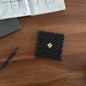 Puede incluir: Un posavasos cuadrado hecho de pequeñas cuentas negras con un diseño central de cuatro cuentas blancas. El posavasos está sobre una superficie de madera oscura, con un bolígrafo y un portátil y un cuaderno parcialmente visibles en el fondo.