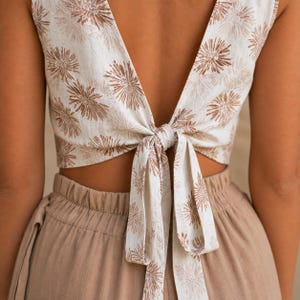 Puede incluir: Un top corto sin mangas con estampado floral marr&oacute;n sobre fondo blanco, dise&ntilde;ado con un escote en V profundo y un cierre de lazo en la espalda. El top se combina con pantalones de color marr&oacute;n claro liso con cintura el&aacute;stica.