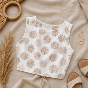Puede incluir: Un top corto sin mangas de color blanco con un patr&oacute;n repetitivo de motivos florales marrones en forma de estrella o diente de le&oacute;n. La prenda tiene un cuello redondo y un dobladillo recto, confeccionada en tela clara con textura.