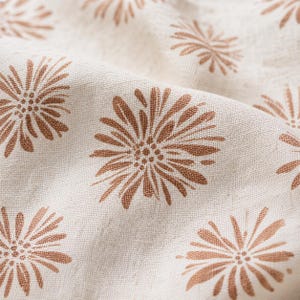 Puede incluir: Una tela de color beige claro con un patr&oacute;n repetitivo de motivos florales estilizados en color &oacute;xido. El tejido presenta una textura visible con pliegues suaves que crean profundidad, resaltando el contraste entre la base clara y el estampado floral.