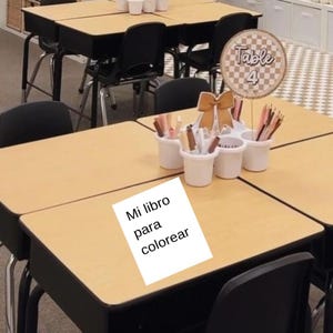 May include: A white paper with the text Mi libro para colorear sits on a light brown desk. Next to it is a white multi-cup organizer filled with colored pencils and a tan bow decoration, topped with a circular sign that reads Table 4.
