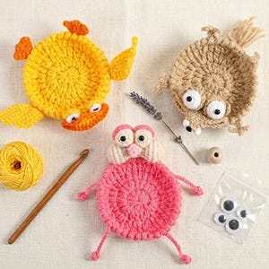 May include: Three crocheted animal coasters in yellow, beige, and pink, accompanied by a ball of yellow yarn, a wooden crochet hook, a sprig of dried lavender, a wooden bead, and a small plastic bag containing four loose plastic googly eyes.