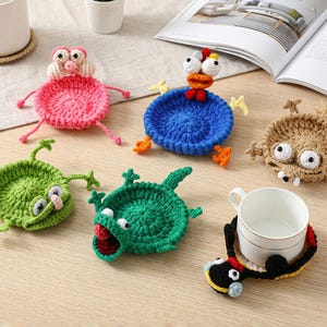 May include: A collection of six colorful crochet coasters shaped like various creatures, including a pink pig, a blue chicken, a tan monster, a light green frog, a dark green alligator, and a black and yellow bird holding a white ceramic mug.