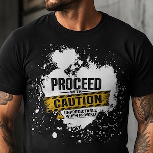Op de afbeelding: Een zwart T-shirt met korte mouwen en een witte verfspat-afbeelding. Het ontwerp bevat de tekst PROCEED WITH CAUTION in zwart en geel, met de aanvullende tekst UNPREDICTABLE WHEN PROVOKED en een geel waarschuwingsicoon in de vorm van een driehoek.