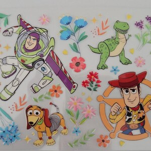 Puede incluir: Una hoja de pegatinas transparente con varios personajes de Toy Story, incluidos Buzz Lightyear, Woody, Jessie, el Sr. Patata, Rex, Slinky Dog, Perdig&oacute;n y Hamm, intercalados con coloridas ilustraciones florales y peque&ntilde;os detalles de estrellas amarillas.