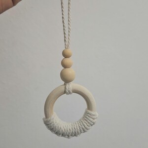 May include: A hanging ornament featuring a smooth wooden ring partially wrapped in off-white cotton cord. Above the ring are three graduated wooden beads strung on a matching cotton cord, which extends upward to form a loop for hanging purposes.