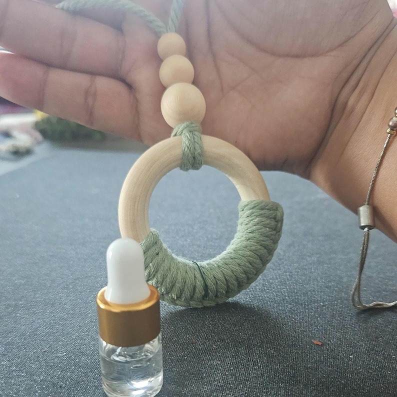 May include: A light green macrame car air freshener featuring a circular wooden ring partially wrapped in sage green cord, accented with three natural wooden beads on a hanging string, paired with a small clear glass bottle with a white dropper.