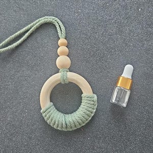 Puede incluir: Un adorno de anillo de macramé en color verde salvia con tres cuentas de madera natural y un lazo de cordón verde salvia para colgar, acompañado de un pequeño frasco de vidrio transparente con cuello dorado y gotero blanco.