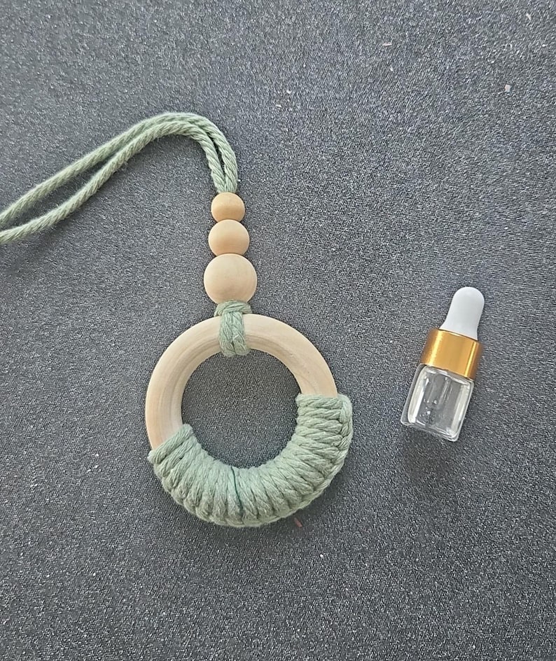 May include: A sage green macrame ring ornament featuring three natural wooden beads and a sage green cord loop for hanging, accompanied by a small clear glass vial with a gold collar and a white dropper top for essential oil application.