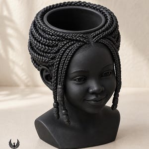 May include: A matte black planter shaped like a human head with detailed braided hair. The hair features intricate cornrows and hanging braids adorned with small cylindrical beads. The top of the head is open for planting. The logo PHOENIX STUDIO is visible.