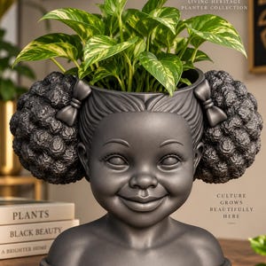 May include: A dark gray bust-shaped planter featuring a smiling face with textured hair styled in two large puffs secured with bows. The planter is labeled Zahara AP-004 Living Heritage Planter Collection and includes the text Culture Grows Beautifully Here.