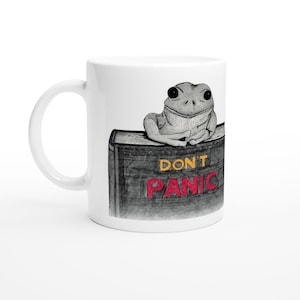 May include: A white ceramic mug featuring a black and white illustration of a frog resting on top of a dark book. The book cover displays the text DON'T in yellow and PANIC in red. The mug has a curved handle on the left side and a smooth finish.