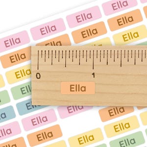 May include: A sheet of rectangular adhesive labels in pastel pink, orange, yellow, green, and blue, each printed with the name Ella in a dark brown font. A wooden ruler is placed over the labels, showing one label is 0.5 inches in width.