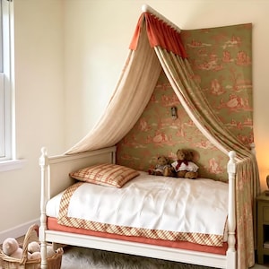 May include: A white wooden bed frame with a canopy featuring a green and orange toile fabric backdrop. The bed is dressed with a white duvet, a plaid pillow, and a plaid bed skirt. Two teddy bears sit on the bed, and a wicker basket sits nearby.