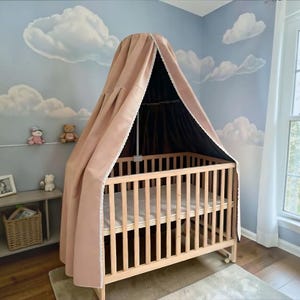 May include: A wooden crib with vertical slats and a mattress, featuring a tall, draped canopy made of solid light brown fabric with white lace trim along the edges. The canopy is supported by a central metal rod attached to the crib frame.