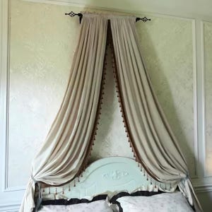 May include: A pair of beige curtains suspended from a black metal rod with decorative finials. The curtains feature a dark brown trim along the inner edges, adorned with small hanging tassels that drape over a light-colored headboard.