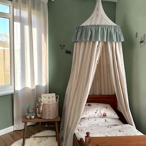May include: A fabric bed canopy featuring vertical stripes in beige and white, topped with a sage green ruffled trim. The canopy hangs over a wooden bed frame with white bedding printed with small forest-themed icons, including trees and acorns.