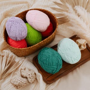 May include: A collection of yarn balls in various colours including lavender, light pink, bright red, lime green, teal, and mint green. The yarn balls are arranged inside a woven brown basket and on a rectangular wooden tray alongside textured stones.
