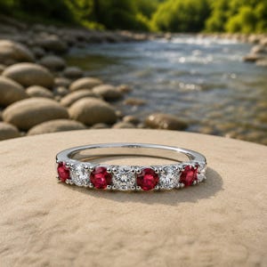 May include: A silver-toned band ring featuring an alternating pattern of round-cut red rubies and clear white diamonds set in a row. The metal band has a polished finish and holds seven stones in total, creating a continuous sparkling design.