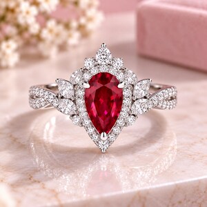 Puede incluir: Un anillo de tono plateado con una gran piedra roja en forma de pera en el centro, rodeada por un halo de pequeñas piedras blancas redondas. Piedras blancas adicionales de corte marquesa forman un patrón floral a lo largo de la banda trenzada.