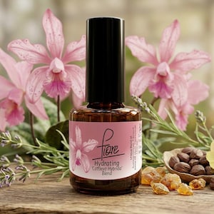 Puede incluir: Un frasco de vidrio marrón con tapa negra y una etiqueta rosa con el texto 'fiore Hydrating Cattleya Hybrida Blend'. El frasco está rodeado de flores de orquídea rosa, ramas de lavanda, un pequeño cuenco de nueces marrones y trozos de resina.