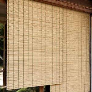 May include: A window blind constructed from thin, light tan horizontal slats held together by vertical green thread. The blind is mounted within a dark wooden frame, featuring a layered design where one section hangs lower than the other.