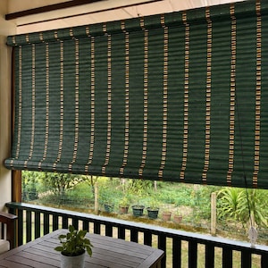 May include: A roll-up bamboo shade featuring a dark forest green finish with vertical brown accents. The shade is mounted within a wooden frame, displaying a textured, slatted construction with a thin pull cord hanging down on the right side.