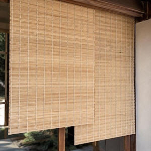 May include: A pair of light brown bamboo roll-up window shades featuring thin horizontal slats held together by vertical white cord stitching. The shades are mounted to a dark wooden frame and hang over a window, providing partial light filtration.