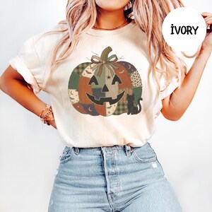 May include: An ivory t-shirt featuring a large graphic of a patchwork pumpkin with a bow on top. The pumpkin design includes various patterns like plaid, leopard print, and moons, with a black cat silhouette and the text IVORY in a circle.