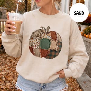 May include: A sand-colored crewneck sweatshirt featuring a central graphic of a pumpkin constructed from various fabric patches, including leopard print, plaid, and autumn leaf patterns, with frayed edges and a small acorn accent at the top.