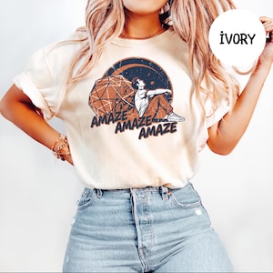 May include: An ivory short-sleeve t-shirt featuring a graphic print of a person sitting next to a geometric sphere against a dark circular background. The text "AMAZE" is printed three times in a stacked, staggered layout below the illustration.