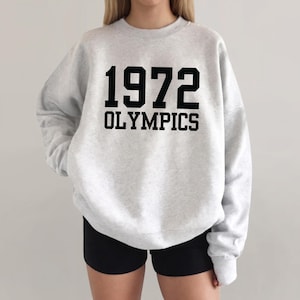 May include: A light gray crewneck sweatshirt featuring the black text 1972 OLYMPICS printed on the chest. The garment has long sleeves, a ribbed neckline, and a relaxed fit, paired with black shorts.