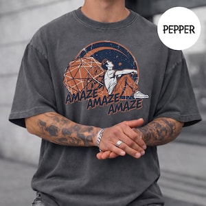 May include: A charcoal grey t-shirt featuring a graphic print of a person sitting next to a large geometric icosahedron shape under a crescent moon. The text "AMAZE" is printed three times in a dark blue font below the illustration. The shirt color is labeled as PEPPER.
